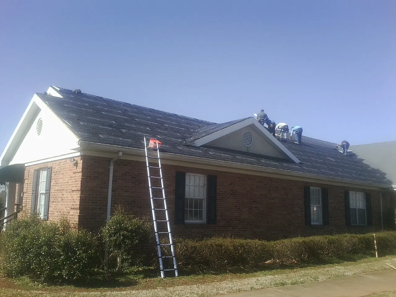 Crew tearing shingles off a brick ranch roof with a ladder set — Metal Roof Repair work by our crew in Albion